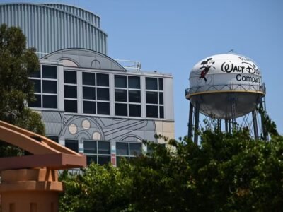 Un château d'eau peint avec le logo de la Walt Disney Company et Mickey Mouse se trouve à côté d'un bâtiment dont l'art ressemble à un appareil photo d'époque. Des arbres verts masquent partiellement le bas de l'image sous un ciel bleu clair, évoquant la magie classique de Disney IA.